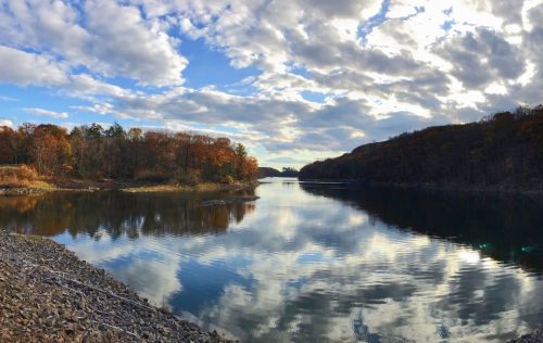 A serene lake surrounded by autumn trees, reflecting clouds and colors in the water under a partly cloudy sky.