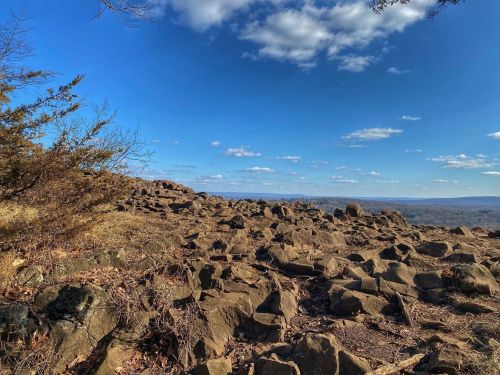 Rocky landscape under a clear blue sky, with scattered clouds and distant hills visible in the background.