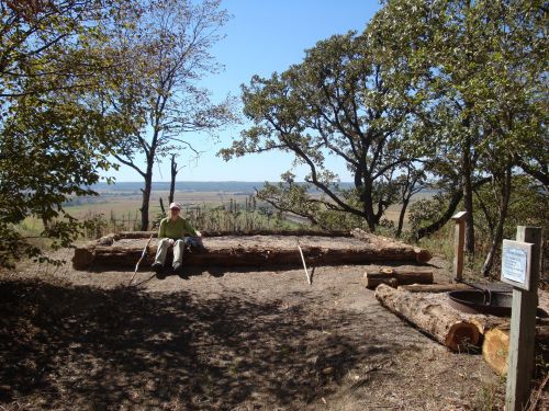 A person sits on a log in a scenic outdoor area with trees and a view of rolling hills in the background.