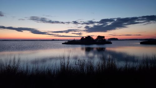 A serene sunset over a calm lake, with silhouettes of trees and an island reflected in the water.
