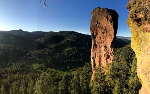 A towering rock formation stands against a backdrop of lush green hills and a clear blue sky.