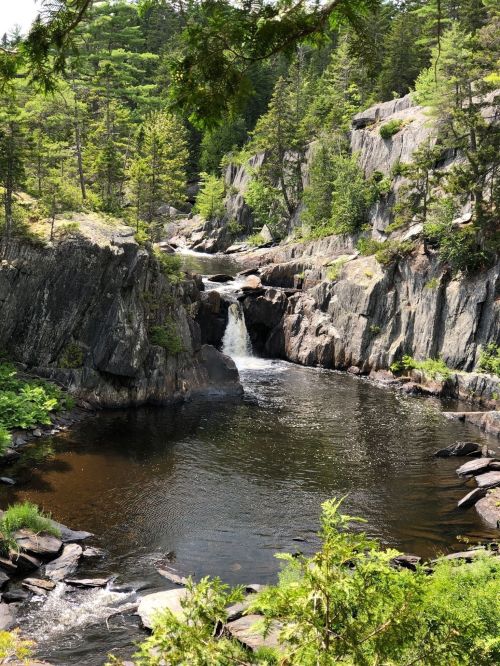 A serene waterfall cascades into a dark pool, surrounded by rocky cliffs and lush green trees.
