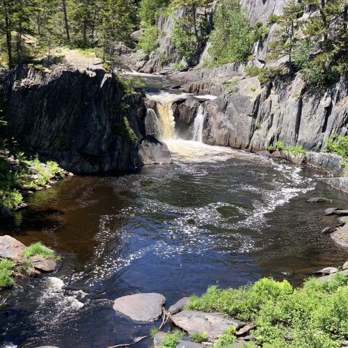 A serene waterfall cascades into a dark river, surrounded by rocky cliffs and lush greenery.