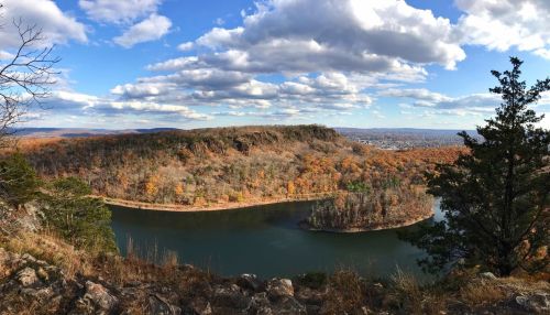 A panoramic view of a river winding through autumn-colored hills under a blue sky with fluffy clouds.