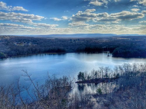 A serene lake surrounded by trees and hills under a blue sky with fluffy clouds.
