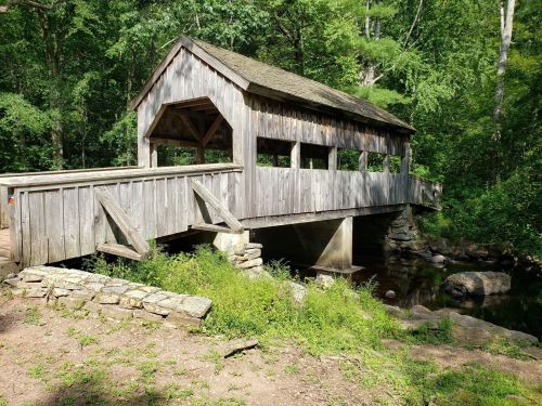 A wooden covered bridge spans a small stream, surrounded by lush greenery and trees.
