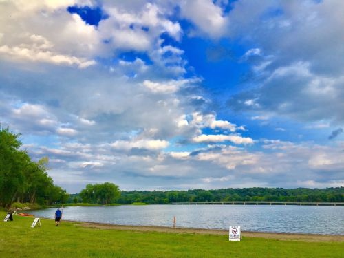 A serene river scene with a person walking along the shore, under a blue sky with clouds and green trees in the background.