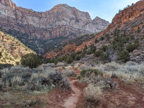 A winding dirt path through a rugged canyon landscape with rocky cliffs and sparse vegetation under a clear blue sky.