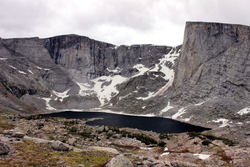 A rugged mountain landscape featuring a dark lake surrounded by rocky cliffs and patches of snow under a cloudy sky.