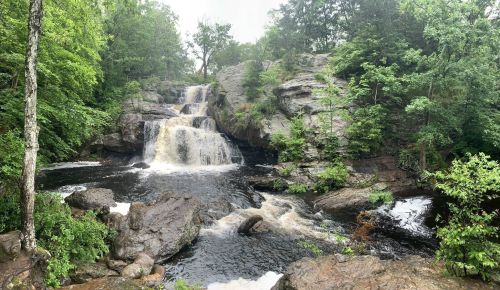 A serene waterfall cascades over rocks, surrounded by lush green trees and a tranquil river.