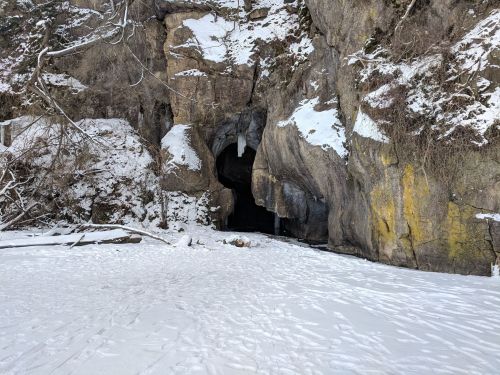A snowy landscape featuring a large rock cave entrance surrounded by rocky cliffs and sparse vegetation.