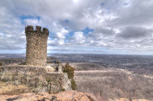 A stone tower atop a rocky hill overlooks a valley with trees and a cloudy sky in the background.
