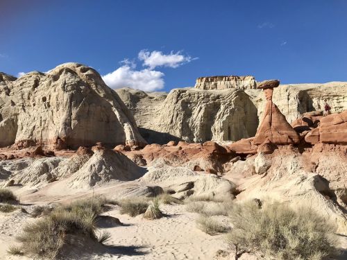 A desert landscape featuring unique rock formations, sandy terrain, and a clear blue sky with scattered clouds.