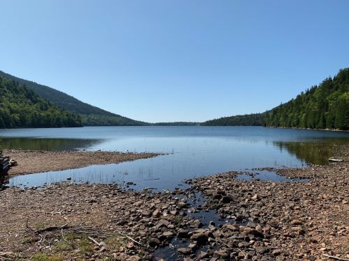 A serene lake surrounded by green mountains under a clear blue sky, with rocky shores and calm water.