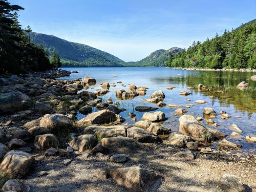 A serene lake surrounded by mountains, with rocky shores and clear blue skies reflecting on the water's surface.