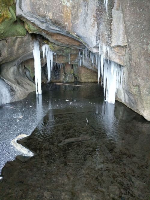 A rocky cave with icicles hanging from the ceiling, reflecting in a still, dark water pool below.