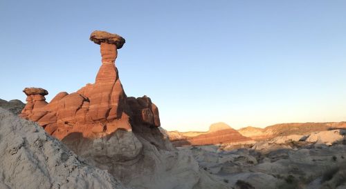 A unique rock formation with a large, flat top stands amidst colorful desert landscape under a clear blue sky.