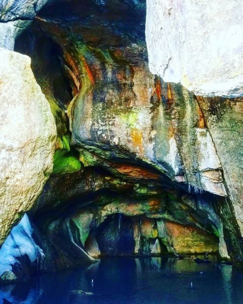 A rocky cave entrance with colorful mineral deposits and a small pool of water inside.