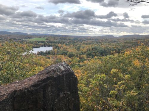 A scenic view of a valley with a river, surrounded by autumn foliage and cloudy skies. A large rock is in the foreground.