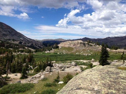 A scenic mountain landscape with rocky terrain, green meadows, and a blue sky dotted with clouds.