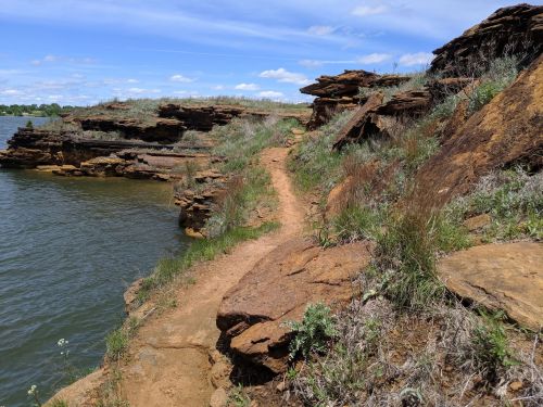 A rocky shoreline with a narrow dirt path, surrounded by grass and water under a blue sky with scattered clouds.