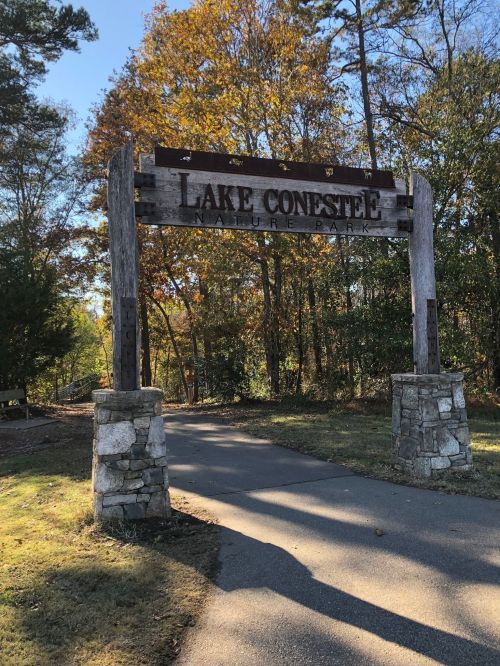 Sign at the entrance of Lake Conestee Nature Park, surrounded by trees and a clear blue sky.