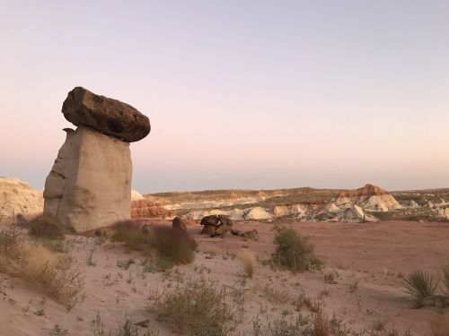 A large rock balanced on a stone pillar in a desert landscape at dusk, with soft pastel colors in the sky.