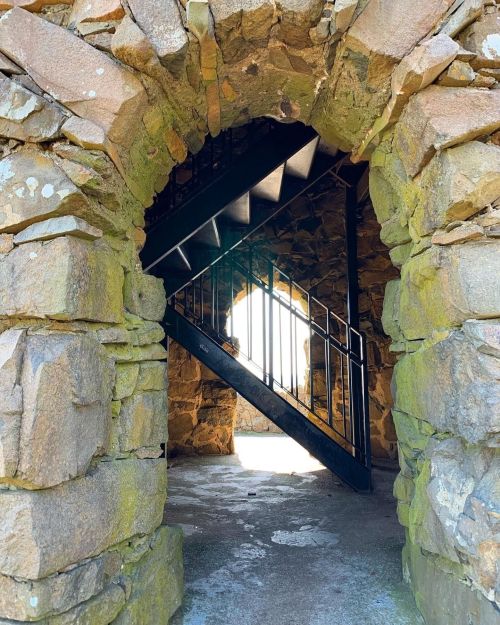 A stone archway frames a black spiral staircase leading upward, with light streaming through an opening above.