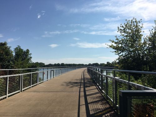 A paved path along a river, bordered by greenery and a metal railing, under a clear blue sky.
