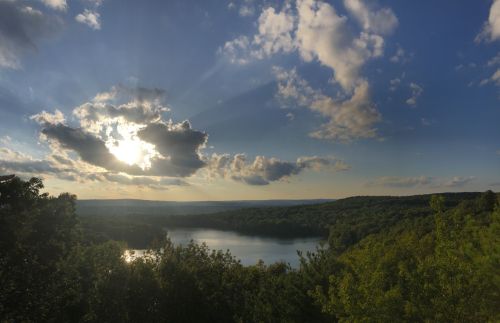 A serene landscape featuring a lake surrounded by lush greenery under a dramatic sky with rays of sunlight breaking through clouds.