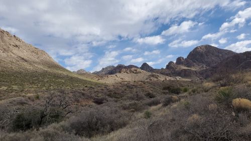 A scenic view of rugged mountains under a partly cloudy sky, with sparse vegetation in the foreground.