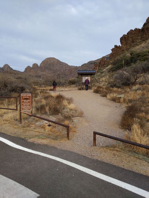 Two people stand near a trailhead sign in a rocky, desert landscape with sparse vegetation and mountains in the background.