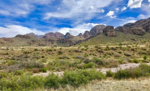 A scenic view of rugged mountains under a blue sky with scattered clouds, surrounded by green shrubs and grassland.