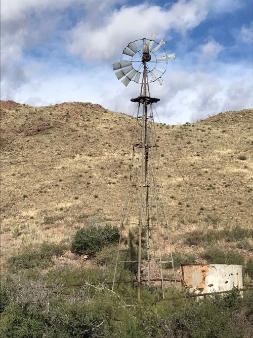 A weathered windmill stands on a grassy hillside under a partly cloudy sky.