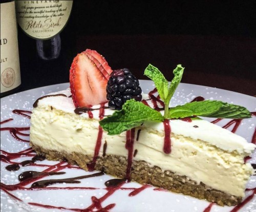 A slice of cheesecake topped with strawberries, blackberries, mint, and a drizzle of berry sauce on a white plate.