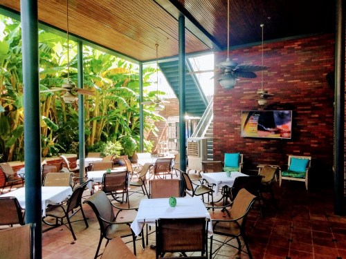 A spacious outdoor dining area with tables, chairs, and tropical plants, featuring a TV and ceiling fans.