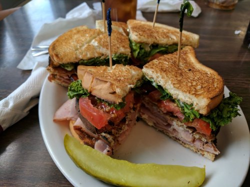A stacked sandwich with layers of meat, lettuce, and tomato, served with a pickle on the side.