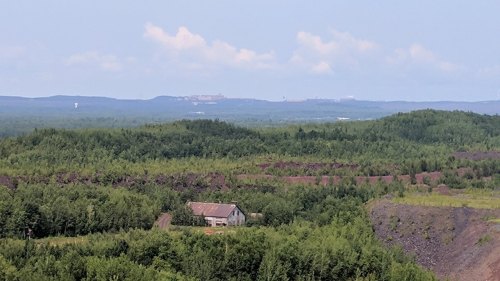 A panoramic view of lush green hills with a distant industrial site and a small building in the foreground.