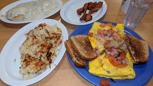 A hearty breakfast plate featuring an omelet with veggies, hash browns, toast, bacon, and a side of biscuits and gravy.