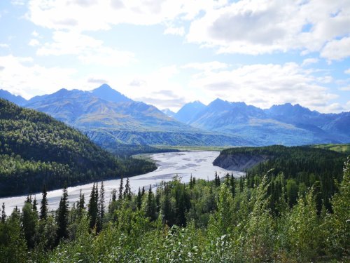 A scenic view of a river winding through a lush valley, surrounded by mountains and a blue sky with scattered clouds.