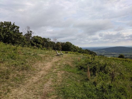 Two wooden chairs sit on a grassy hilltop, overlooking a scenic valley under a cloudy sky.