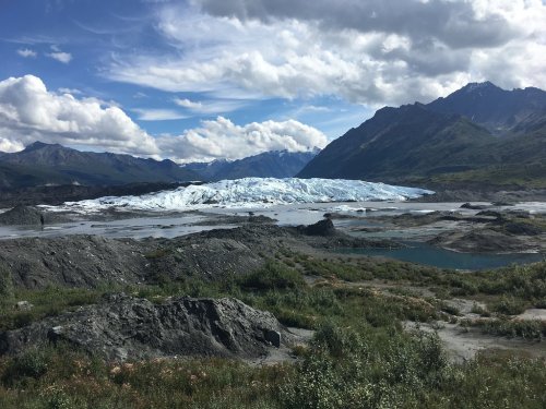 A vast glacier surrounded by mountains under a partly cloudy sky, with rocky terrain and a small body of water in the foreground.