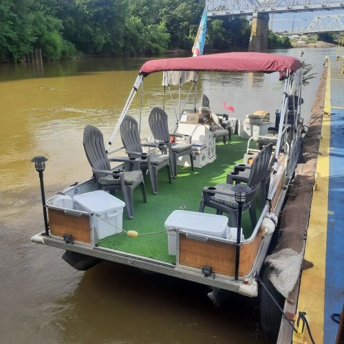 A pontoon boat with chairs and a green carpet, docked on a river surrounded by trees.