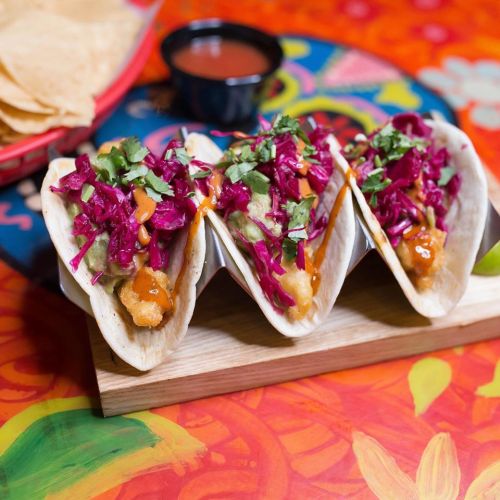 Three tacos filled with colorful slaw and sauce, served on a wooden board with a side of salsa and tortilla chips.