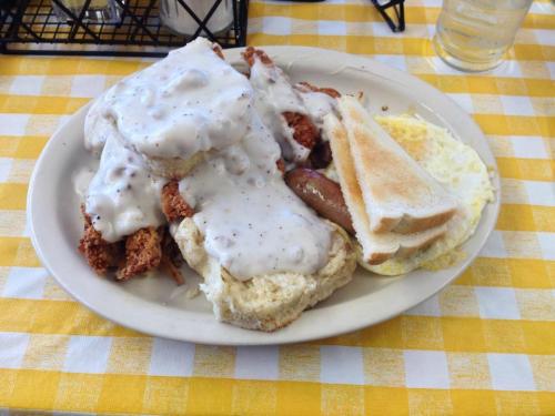 A plate of fried chicken topped with creamy gravy, biscuits, sausage, and toast, served on a checkered tablecloth.