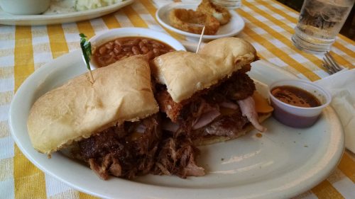 A plate with a pulled pork sandwich, baked beans, and a side of sauce, all on a checkered tablecloth.