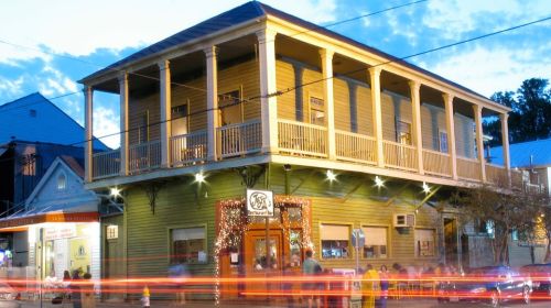 Historic building with a wraparound porch, illuminated at dusk, surrounded by shops and streetlights.