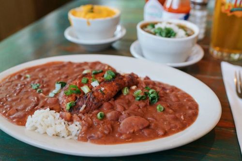 A plate of rice and red beans topped with grilled chicken, garnished with green onions, alongside bowls of soup and cheese dip.