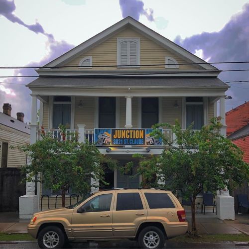 A two-story yellow house with a large sign reading "JUNCTION 3021 ST. CLAUDE" and a parked beige SUV in front.