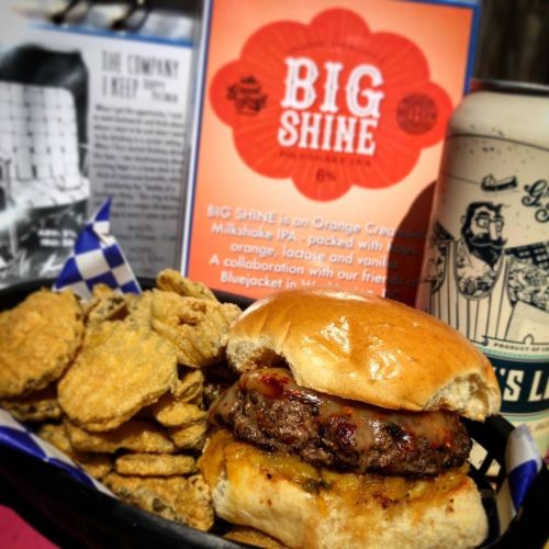 A juicy burger with melted cheese, served with crispy pickles and a can of Big Shine beer in the background.
